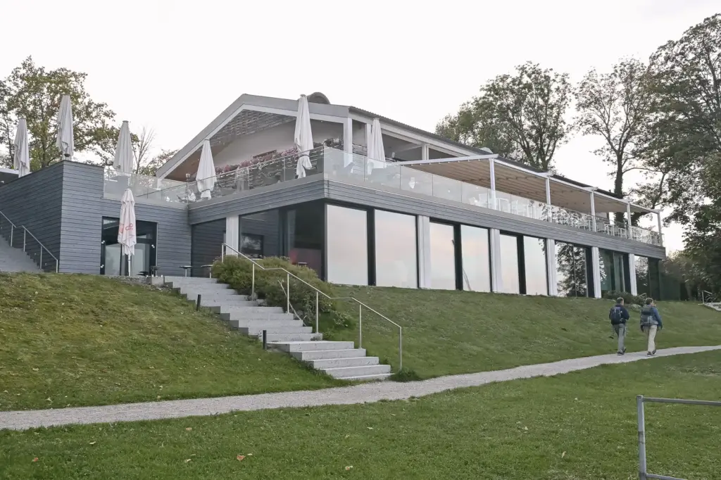 Das Marina Hotel am Yachthafen Bernried von der Seeseite, mit Terrasse über großen Glasfenstern und einer Steintreppe über der Wiese