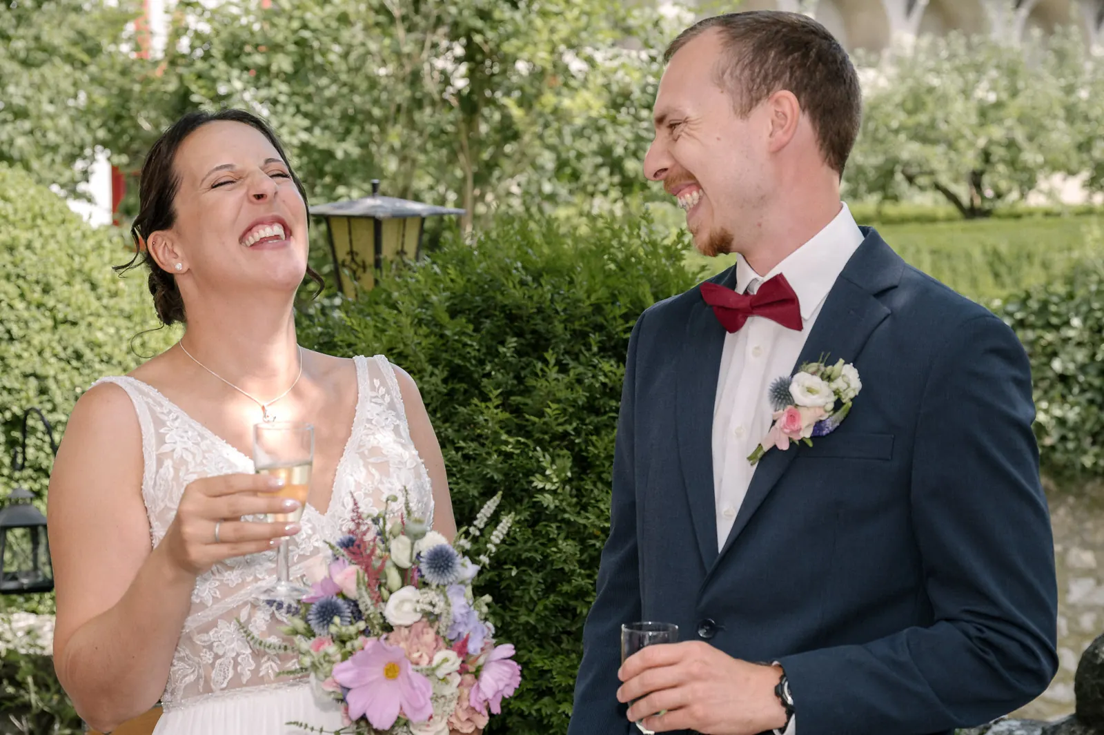 Braut mit Sektglas und Brautstrauß und Bräutigam lachen herzhaft