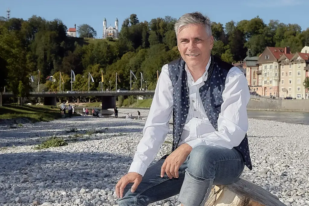 Harry Wolfsbauer sitzt in Trachtenweste und Jeans auf einem Baumstamm an der Isar in Bad Tölz, dahinter eine Brücke und Häuser