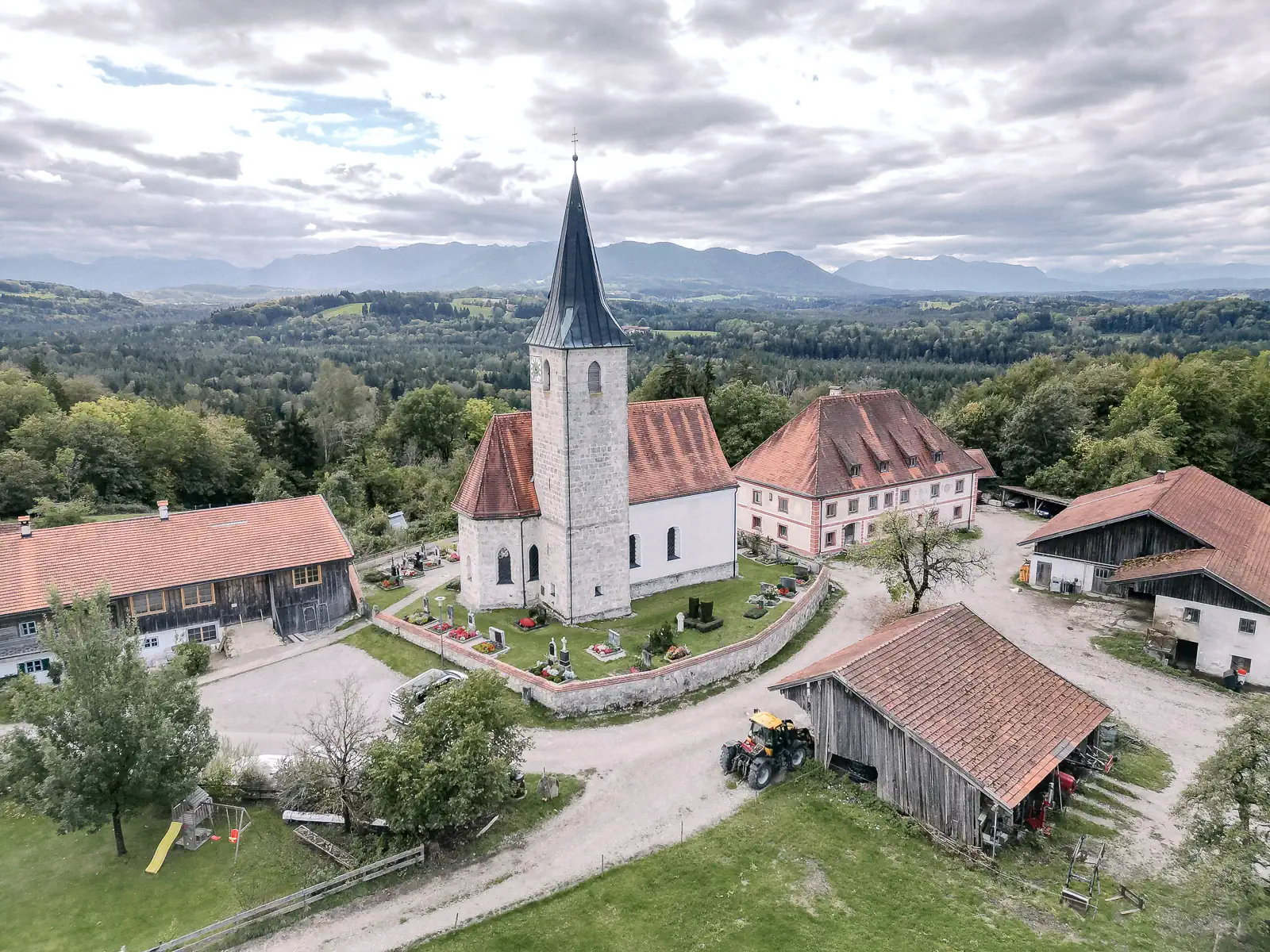 Luftaufnahme der Kirche St. Valentin in Hechenberg mit Friedhof, umgeben von Bauernhöfen, dahinter Wald und Berge