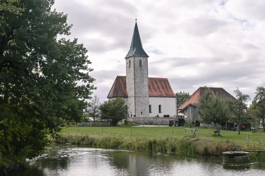 Die Kirche St. Valentin in Hechenberg mit spitzem Turm, davor eine Wiese und ein Weiher