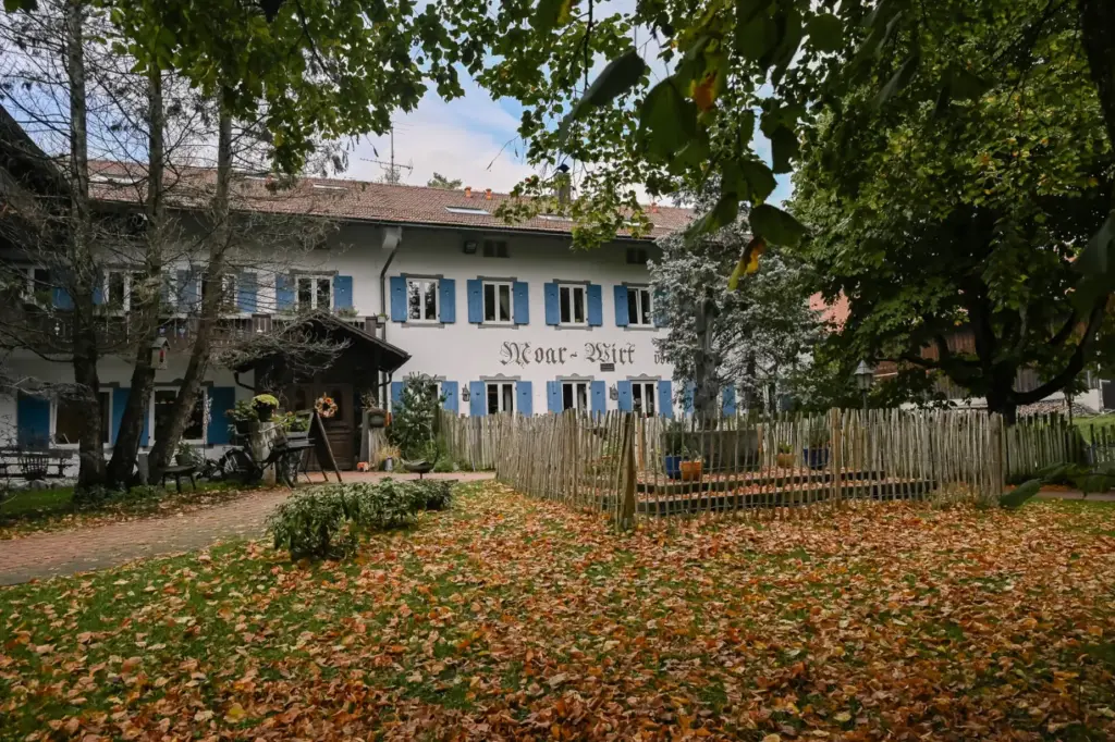 Die Fassade des Moarwirts in Hechenberg mit blauen Fensterläden und dem Schriftzug Moar-Wirt, davor Herbstlaub auf der Wiese