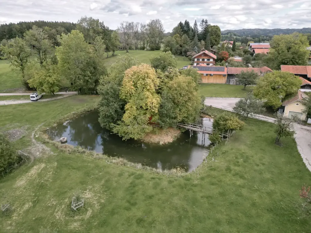 Luftaufnahme eines kleinen Weihers mit einer bewachsenen Insel und einer Holzbrücke in Hechenberg