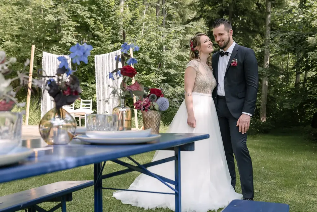 Brautpaar steht auf einer Lichtung neben einem blau gedeckten Biertisch mit Blumen, dahinter Spitzenvorhänge und Wald